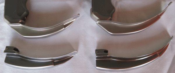 **Imaging Modality:** Clinical photograph of medical instrumentation.

**Device Classification:** Comparison of airway management tools, specifically laryngoscope blades.

**Key Components:** 
The image displays four metallic laryngoscope blades arranged in a 2x2 grid. 
*   **Superior Row:** Two C-MAC videolaryngoscope blades (sizes 3 and 4). These feature integrated digital camera housings and are attached to specialized ergonomic handles. The blade geometry maintains a traditional Macintosh-style curve but with a slimmer profile to accommodate the electronic optical components.
*   **Inferior Row:** Two conventional Macintosh laryngoscope blades (sizes 3 and 4). These are standard direct laryngoscopy blades characterized by a broad, curved spatula and a proximal heel designed for attachment to a standard battery-operated handle.

**Characteristic Visual Features:**
The comparison highlights the structural similarities in curvature (curved blade design for displacement of the tongue into the submandibular space) while illustrating the design modifications required for videolaryngoscopy. The C-MAC blades exhibit a more pronounced proximal housing for the video sensor compared to the thinner, purely mechanical hinge of the standard Macintosh blades.

**Clinical Context:** These devices are utilized for endotracheal intubation and glottic visualization during anesthesia and emergency airway management.