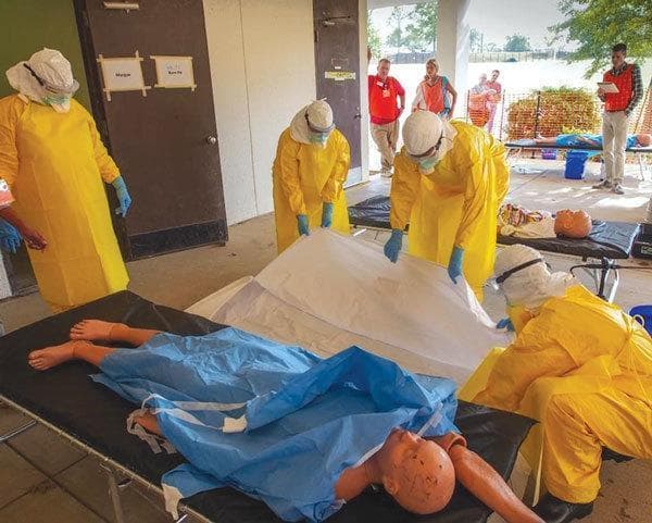 A clinical photograph of a medical training exercise at a mock Ebola Treatment Unit (ETU). The image shows healthcare trainees wearing full Personal Protective Equipment (PPE) ensembles designed for high-consequence viral hemorrhagic fevers. The PPE consists of yellow fluid-resistant coveralls, white integrated hoods, eye protection (goggles or face shields), N95 respirators, blue nitrile gloves, and black rubber gum boots. The trainees are practicing post-mortem care and infection prevention and control (IPC) protocols by preparing a simulated deceased patient (a medical mannequin) for placement into a white body bag. The mannequin is partially covered by a blue surgical sheet, with visible markings on the face representing clinical signs of disease. The background shows a controlled environment with clearly marked entry/exit points, illustrating the unidirectional flow and triage protocols used in field hospitals. This visual serves as an educational resource for biohazard safety training, donning and doffing procedures, and managing infectious disease outbreaks in resource-limited settings.