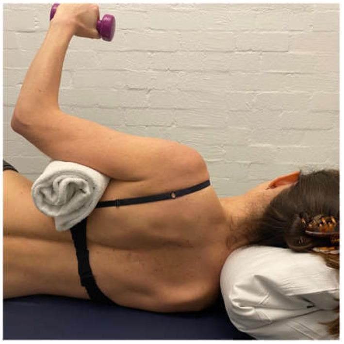 This clinical photograph demonstrates a side-lying external rotation (ER) exercise, a key rehabilitation technique for glenohumeral joint stabilization and rotator cuff strengthening. The patient is positioned in a side-lying posture with a pillow supporting the head. The superior arm is positioned in approximately 0 to 20 degrees of abduction, maintained by a rolled white towel bolster placed between the thorax and the medial aspect of the humerus to optimize subacromial space and rotator cuff recruitment. The elbow is flexed to 90 degrees, and the patient is performing active external rotation against gravity while holding a small purple dumbbell. A black elastic strap is visible across the mid-back, likely utilized for postural cues or scapular stabilization. This exercise is specifically indicated for patients with multidirectional instability (MDI) or posterior humeral head translation, as it aims to strengthen the posterior musculature (infraspinatus, teres minor, and posterior deltoid) and improve scapular motor control.