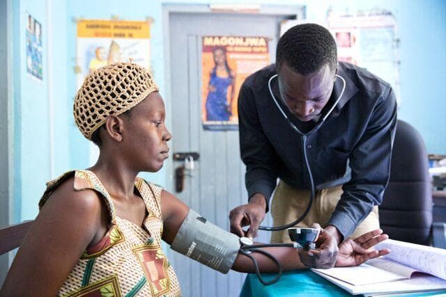 A clinical photograph depicting a healthcare provider, identified as a clinical officer, performing a physical assessment on a female patient in a community health facility. The primary activity shown is the measurement of arterial blood pressure using a manual aneroid sphygmomanometer and a stethoscope. The provider is seen adjusting the inflation bulb and observing the manometer gauge while the cuff is secured around the patient's left upper arm. The setting is a clinical workspace featuring health education posters (one labeled 'MAGONJWA') and medical documentation. This visual illustrates routine preoperative screening or vital sign monitoring within the context of reproductive health services, specifically related to assessing eligibility for surgical contraception such as tubal ligation. The image emphasizes clinical interactions and the use of basic diagnostic devices in primary care or rural healthcare settings.