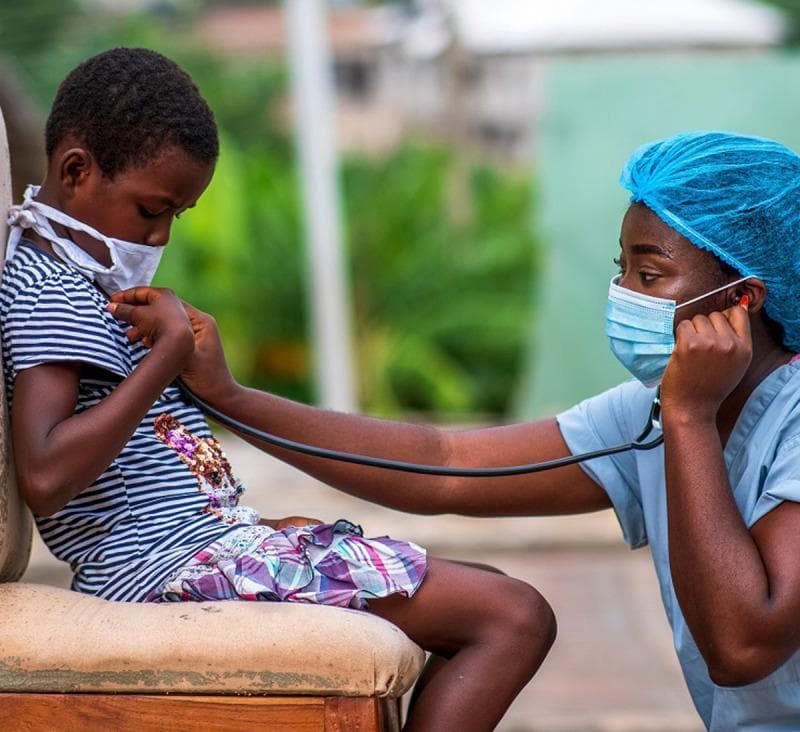 This clinical photograph depicts a pediatric physical examination conducted in an outdoor or informal healthcare setting. A healthcare professional, wearing blue personal protective equipment (PPE) including a surgical cap, face mask, and scrubs, is performing auscultation of a young child's chest using a stethoscope. The pediatric patient is also wearing a protective white face mask, indicating adherence to infection control protocols during a medical consultation. The child is actively participating by helping to position the stethoscope diaphragm against their chest. This image illustrates concepts of primary care, pediatric cardiology or pulmonology, and global health outreach. The presence of comprehensive PPE for both the provider and patient highlights the importance of respiratory hygiene and airborne pathogen mitigation in clinical practice.
