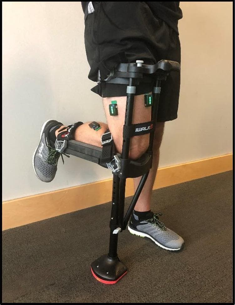 A clinical photograph illustrating a hands-free single crutch (HFSC), specifically the iWALKFree model, fitted to a user's dominant lower extremity. The device is designed for non-weight-bearing restrictions, showing the lower leg bent at a 90-degree angle at the knee and resting horizontally on a padded knee plate. The HFSC is secured with adjustable straps around the upper thigh and lower leg. Also visible are three Trigno Avanti wireless Bluetooth electromyography (EMG) sensors attached to the skin using adhesive pads. These sensors are strategically placed over the bellies of the rectus femoris, vastus lateralis, and lateral gastrocnemius muscles to monitor muscle activity and engagement during gait. The image demonstrates the clinical application of ambulatory aids combined with wearable sensor technology used to assess muscle atrophy prevention and gait analysis in orthopedic rehabilitation.