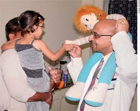 A clinical photograph illustrating a pediatric ophthalmic examination technique. A male physician, wearing a white coat, glasses, and a tie, is shown interacting with a young girl who is wearing corrective spectacles and being held by a parent. The clinician has a large, colorful plush doll with orange hair draped over his shoulders, serving as a distraction tool to build rapport and decrease patient anxiety during the physical assessment. He is gently holding the child's hand while engaging in playful interaction. The background shows clinical equipment, including a slit lamp biomicroscope, indicating an eye clinic setting. This image demonstrates the 'playful examination' principle in pediatrics, which is essential for ensuring cooperation during diagnostic procedures such as the red reflex test or visual acuity assessments in young children. The educational focus is on non-pharmacological behavioral management and communication strategies to facilitate a successful medical evaluation in a pediatric population.