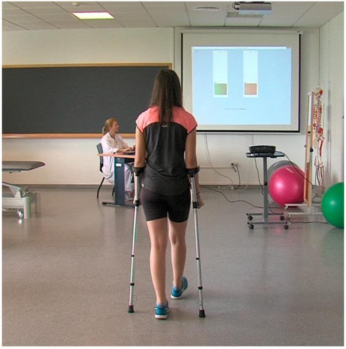 This clinical photograph illustrates a physical therapy and gait rehabilitation setting focusing on assistive device training. A female patient is seen from behind, ambulating with a pair of adjustable forearm crutches. These crutches are equipped with integrated force sensors designed to monitor partial weight-bearing (PWB) compliance. In the background, a large projection screen provides real-time visual biofeedback through two vertical bar charts; the left bar is green and the right is orange, representing the load distribution for each crutch. A healthcare professional monitors the session from a workstation. The setting includes common rehabilitation equipment such as an anatomical skeleton model and therapy balls. This setup demonstrates a closed-loop feedback mechanism in gait training, where sensor-based data is used to help patients self-correct their loading technique and adhere to prescribed weight-bearing limits during the recovery of lower limb pathologies.