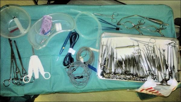 This intraoperative clinical photograph displays a surgical instrument setup on a Mayo stand draped in green sterile fabric, prepared for a thyroid or neck procedure. On the left, a circular basin contains a syringe and needles, while a kidney dish holds gauze and packaged scalpel blades. Below these, long vascular forceps and a three-pronged white self-retaining retractor are visible. The center area contains coiled electrosurgical cords, a blue bovie pen, and suction tubing. The right side features a large, rectangular tray densely packed with specialized surgical tools, including Macindoe scissors, dissection clips, various fine-tipped forceps, and clamps. Additional instruments outside the tray on the upper right include hinged retractors and slender grasping tools. The arrangement illustrates standard operating room protocol for sharp safety, using kidney dishes for transfer and organized trays for instrument management. This visual serves as an educational resource for surgical technology, perioperative nursing, and surgical residency training, emphasizing the organization and nomenclature of head and neck surgical instrumentation.