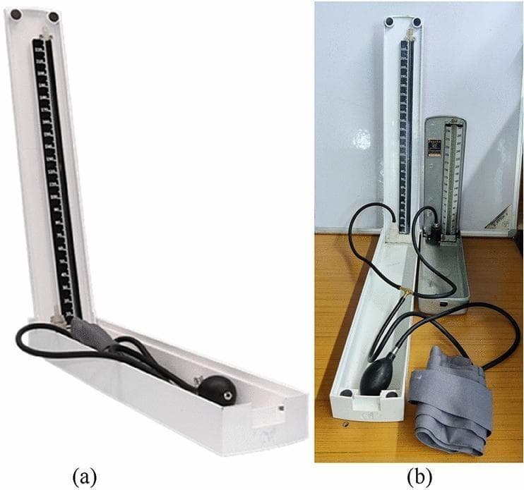 This clinical photograph displays a comparative analysis of sphygmomanometer devices used for blood pressure measurement. Panel (a) shows the 'Merkfree' alpha-prototype, a mercury-free sphygmomanometer featuring a white 3D-printed thermoplastic (PLA) enclosure. The design includes a horizontal base compartment containing a black rubber inflation bulb and tubing, and a vertical measuring scale with high-contrast markings calibrated for mmHg. The device utilizes Galinstan, a non-toxic liquid metal alloy, as the measuring fluid within a glass tube, maintaining the traditional vertical column interface. Panel (b) provides a technical comparison between the Merkfree prototype and a standard Mercurial Sphygmomanometer (MS). Both devices are connected via a T-connector joint to a single inflation bulb and blood pressure cuff (visible in the foreground) to ensure simultaneous pressure application during validation trials. The comparison highlights differences in enclosure materials—white polymer for the prototype versus metallic for the conventional unit—while demonstrating the similar structural logic intended to maintain clinician familiarity and reduce rounding-off errors through improved scale calibration.