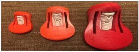 This clinical photograph displays three 3D-printed anatomical models used as cricothyroidotomy trainers, representing infant, child, and adult scales. Arranged from left to right in ascending size, the trainers consist of a molded red or orange-red plastic base housing a white internal anatomical insert. Each insert depicts the larynx and upper trachea, featuring landmarks such as the thyroid cartilage, cricoid cartilage, and tracheal rings. The models highlight developmental differences in airway anatomy, with the adult model on the right showing the most pronounced laryngeal prominence and larger cricothyroid membrane space. These specimens are used in surgical skills labs for emergency airway management training, specifically for practicing the identification of anterior neck landmarks and the performance of surgical cricothyroidotomy across different patient demographics. The models demonstrate the application of 3D printing in creating low-cost, age-specific medical simulation tools for emergency medicine and surgical education.