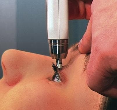A clinical photograph demonstrating the proper application of a Diaton® transpalpebral tonometer for measuring intraocular pressure (IOP). The image depicts the handheld device being applied to a patient in a supine or reclined position. The tonometer is held vertically, perpendicular to the ground, with the tip making contact with the skin of the superior eyelid at the level of the tarsal plate. The patient's eye is partially open, showing the sclera and iris, while the device is positioned above the lash line to avoid direct corneal contact. The clinician's hand is visible, stabilizing the device against the orbital rim or maxilla to ensure accuracy and minimize movement. This method of transpalpebral tonometry is an alternative to Goldmann applanation, as it allows for IOP measurement through the eyelid without anesthesia or contact with the cornea, making it useful in clinical scenarios where corneal contact is contraindicated or difficult.