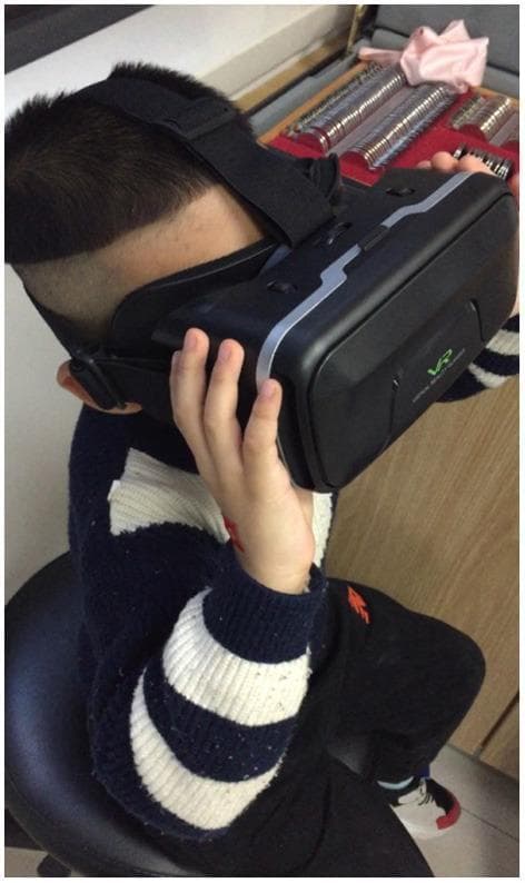 This clinical photograph illustrates a pediatric patient engaged in amblyopia therapy using the EYEBIT system, a digital health intervention based on virtual reality (VR) technology. The patient is seated and wearing a black VR headset with silver-grey accents and a visible 'VR' logo, secured by adjustable elastic head straps. In the background, a trial lens set is visible, typical of an ophthalmology or optometry clinical setting. The system utilizes dichoptic stimulation, where the visual stream is manipulated between eyes—specifically by partially covering the central area of the dominant eye's window to force visual training and stimulation of the central vision in the amblyopic eye. This setup is designed to improve binocular fusion function and visual acuity through active stimulation rather than traditional passive patching. The image demonstrates a modern approach to neuro-rehabilitation in ophthalmology, focusing on binocular visual function and patient compliance in treating refractive or strabismic amblyopia.