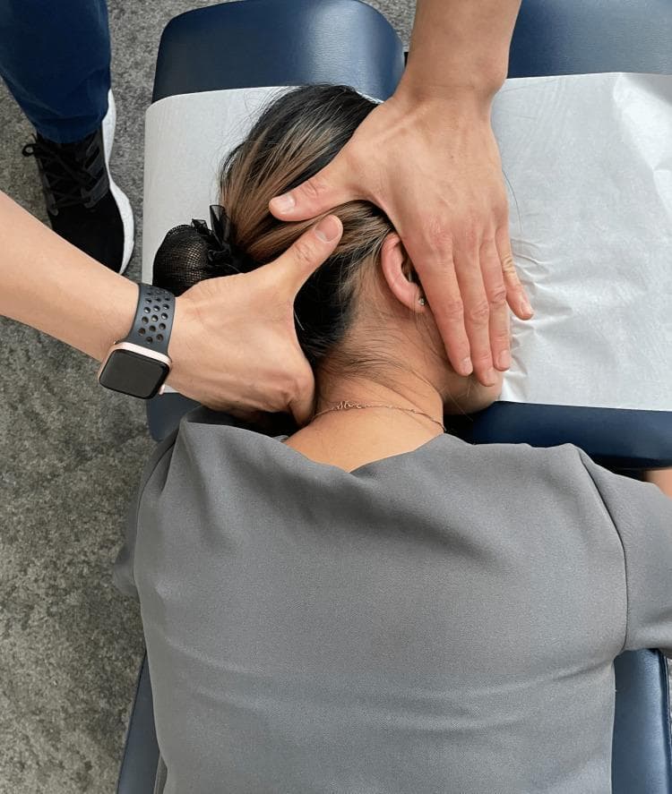 A clinical photograph demonstrating a manual therapy technique identified as prone cervical spinal manipulation. The patient is positioned prone on a chiropractic or osteopathic treatment table with the head resting on a headrest covered by protective paper. The practitioner is shown at the head of the table applying specific hand placements to the cervical region. The left hand provides stabilization of the patient's head on the left side, while the right hand is positioned with the palm over the ear and the index finger contacting the lateral cervical spine. This setup is characteristic of a high-velocity, low-amplitude (HVLA) thrust directed toward the C5/C6 spinal segments. The educational focus is on the biomechanical setup, patient positioning, and contact points necessary for delivering a targeted adjustment to the mid-to-lower cervical spine in a prone position. This image serves as a clinical reference for manual medicine, physical therapy, and chiropractic education.