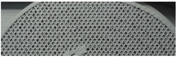 This image displays a macroscopic clinical photograph of a 3D-printed capillary model designed for biomedical sensing applications. The structure features a distinctive honeycomb-like repeating pattern of interconnected, uniform hexagonal or diamond-shaped cells. This porous architectural arrangement is characteristic of synthetic scaffolds or filtration membranes used in medical engineering to mimic natural capillary beds or to facilitate gas exchange. The overall form factor is semi-circular, suggesting a cross-sectional view of a multi-tier cylindrical column. The small, dark openings represent the individual capillary elements, which are densely packed to maximize the surface-area-to-volume ratio. This design is clinically relevant for studies involving scent-based diagnostic sensors or hemodialysis membrane development, where precise flow control and interaction between a gas or liquid sample and a sensing element are required. The material appears uniform, highlighting advancements in precision medical manufacturing for creating complex anatomical analogues.