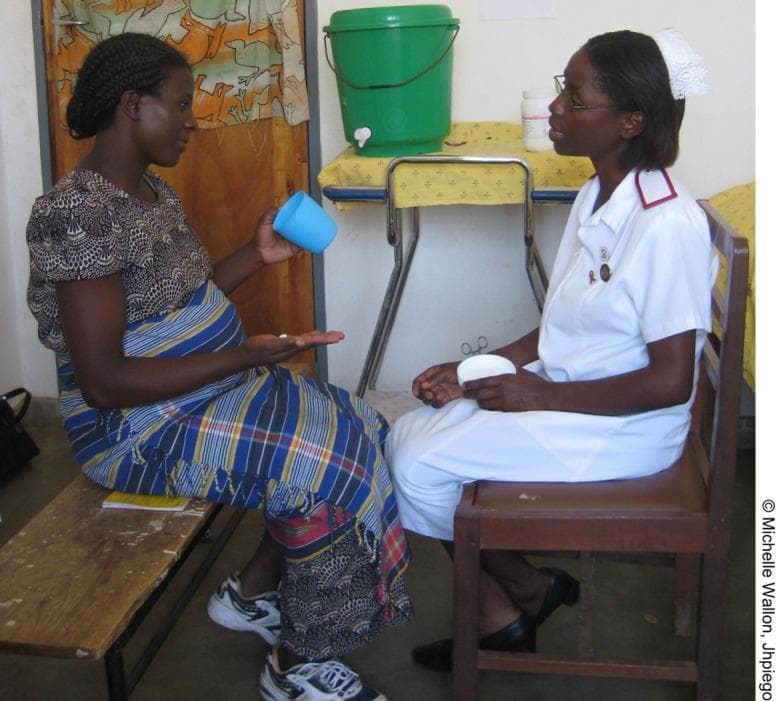 A clinical photograph depicting an antenatal care (ANC) visit in a global health setting, illustrating the administration of Intermittent Preventive Treatment of malaria in pregnancy (IPTp). The image shows a pregnant woman in the third trimester seated on a bench, holding oral medication (pills) in one hand and a blue cup of water in the other, demonstrating Directly Observed Therapy (DOT). Across from her, a healthcare provider in a professional white nurse's uniform with a red cross emblem and nursing cap provides clinical guidance. The setting includes essential clinic supplies, such as a water dispenser and medical documentation on a yellow notepad. This visual serves as an educational example of maternal health interventions, specifically the prevention of Malaria in Pregnancy (MiP) through health system scaling and standardized diagnostic/treatment protocols in resource-limited environments.
