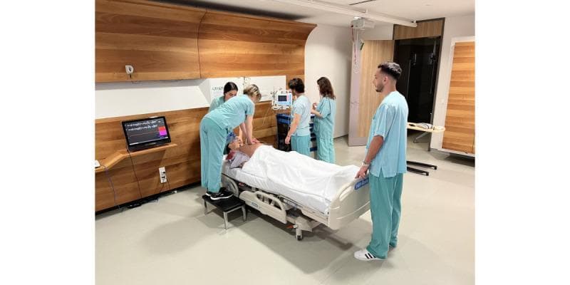 A clinical photograph depicting a high-fidelity medical simulation of an Advanced Life Support (ALS) scenario. The image shows a team of five healthcare students or professionals in light blue scrubs attending to a high-fidelity patient simulator (Apollo Patient Simulator) positioned on a standard adjustable hospital bed. One rescuer is standing on a step stool performing chest compressions on the manikin's exposed chest, while others manage the airway and monitor vital signs. The educational environment is equipped with authentic clinical technology, including a patient monitor displaying multi-parameter waveforms (ECG, SpO2) and a secondary laptop screen showing detailed cardiac rhythm data. The setting is a specialized simulation lab designed to replicate a modern hospital room, used for teaching Crisis Resource Management (CRM) skills, resuscitation algorithms, and technical medical interventions in a controlled, risk-free setting.