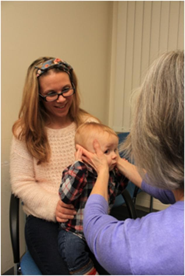 Clinical photograph demonstrating the 'doll's head manoeuvre' (oculocephalic reflex testing) performed on an infant in a pediatric or neuro-ophthalmological setting. The infant is seated on a caregiver's lap, who provides trunk stabilization. A clinical examiner, positioned in front of the infant, gently but firmly holds the child's head with both hands placed laterally over the parietal-temporal regions. The image illustrates the correct positioning for manual head rotation to assess the Vestibulo-Ocular Reflex (VOR). The procedure involves rapid horizontal or vertical head movements while observing the infant's eyes; a normal response is the maintenance of steady gaze or eye movement in the direction opposite to the head rotation. This clinical test is essential for evaluating brainstem function and vestibular system integrity in non-verbal or uncooperative pediatric patients, provided there are no contraindications such as cervical spine instability.