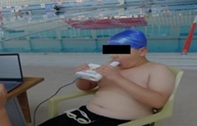 This clinical photograph illustrates the performance of a pulmonary function test, specifically Forced Vital Capacity (FVC) measurement, in an aquatic sports setting. A young male subject, wearing a blue swimming cap and dark shorts, is seated while performing spirometry. He is using a handheld digital spirometer (Pocket Spiro USB-100), gripping the white device with both hands. The subject’s lips are tightly sealed around a disposable cylindrical mouthpiece, and a nose clip is applied to ensure isolated oral expiration. A data cable connects the spirometer to a laptop on a nearby table, indicating real-time data acquisition and flow-volume loop analysis. The background shows a swimming pool environment, suggesting clinical research into the respiratory physiology of swimmers or the impact of aquatic training on lung volumes. This image serves as an educational example of portable spirometry application and proper patient positioning for diagnostic respiratory maneuvers outside a traditional lab setting.