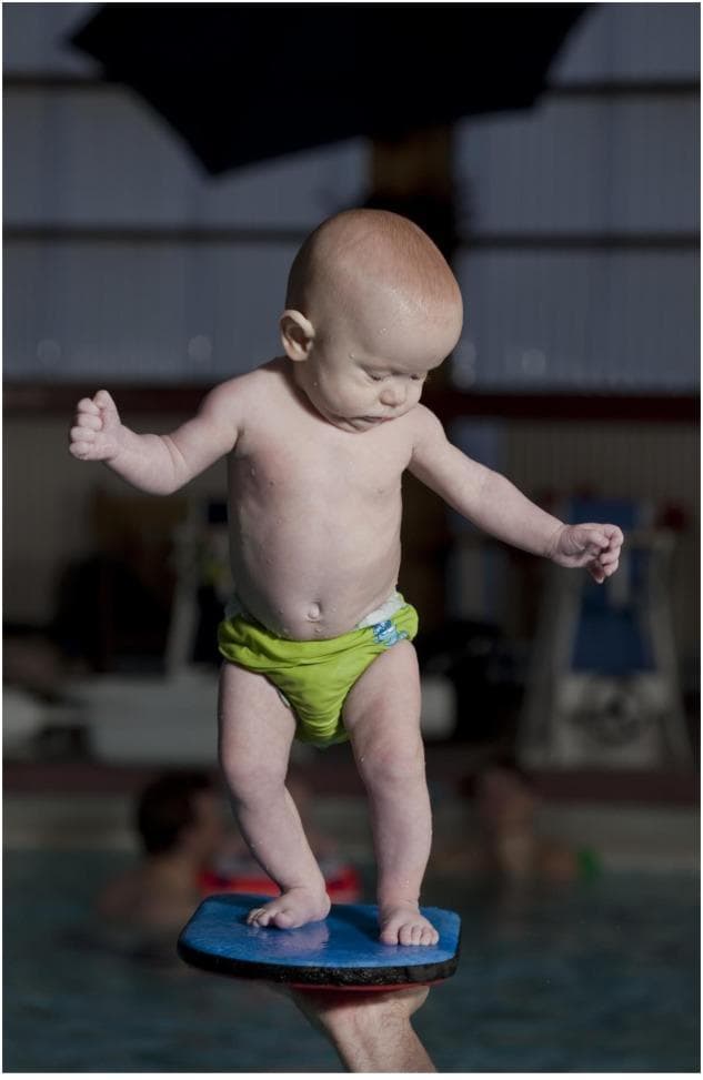 Clinical photograph of a 4.8-month-old infant demonstrating early motor development through an independent standing task. The infant is positioned on a buoyant blue corkboard held by an instructor over a pool of water, providing a moderately stable but dynamic base of support. Visually, the infant exhibits high-guard arm positioning—abducted and extended—to maintain balance, characteristic of early postural control development. The lower extremities are slightly flexed at the knees and hips, with a neutral foot placement to stabilize the center of gravity. The infant's gaze is directed downward, indicating visual engagement with the supporting surface to assist proprioceptive feedback. This image is used in pediatric developmental assessments to illustrate gross motor milestones, specifically the emergence of weight-bearing and trunk stability in the pre-walking phase. The setting and skin appearance suggest an aquatic therapy or infant swimming context, highlighting the role of environmental factors and external support in eliciting advanced motor responses.