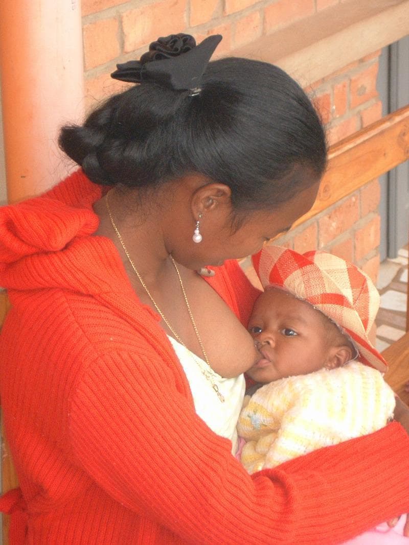 A clinical photograph depicting a mother breastfeeding an infant in a community setting. The image illustrates proper positioning and latch during lactation. The infant is held close in the mother's arms, positioned at the breast, demonstrating active suckling. The mother's posture shows her leaning slightly toward the infant to facilitate contact. The scene provides clinical context for maternal and child health, particularly regarding breastfeeding practices, infant nutrition, and the physiological process of lactation. The educational focus is on postnatal care, maternal-infant bonding, and the promotion of exclusive breastfeeding in global health contexts. Key elements include the physical interaction required for successful feeding and the anatomical alignment of the infant during the breastfeeding process.
