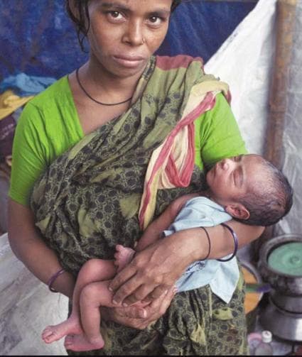 Clinical photograph of a mother holding a newborn infant in a domestic or field setting, representative of maternal and child health studies in South Asian regions like Dhaka, Bangladesh. The mother is shown in a three-quarter view, wearing traditional attire including a green blouse and a floral-patterned shawl. The newborn is positioned in the mother's arms in a resting, supine-to-sidelying posture. The infant has light brown skin, visible scalp hair, and appears stable with no immediate signs of acute respiratory distress or visible cutaneous lesions. From an educational perspective, this image illustrates neonatal care, the postpartum period, and the socio-economic determinants of health in developing urban environments. It serves as a visual reference for community health outreach, breastfeeding support, and pediatric assessment in global health contexts.
