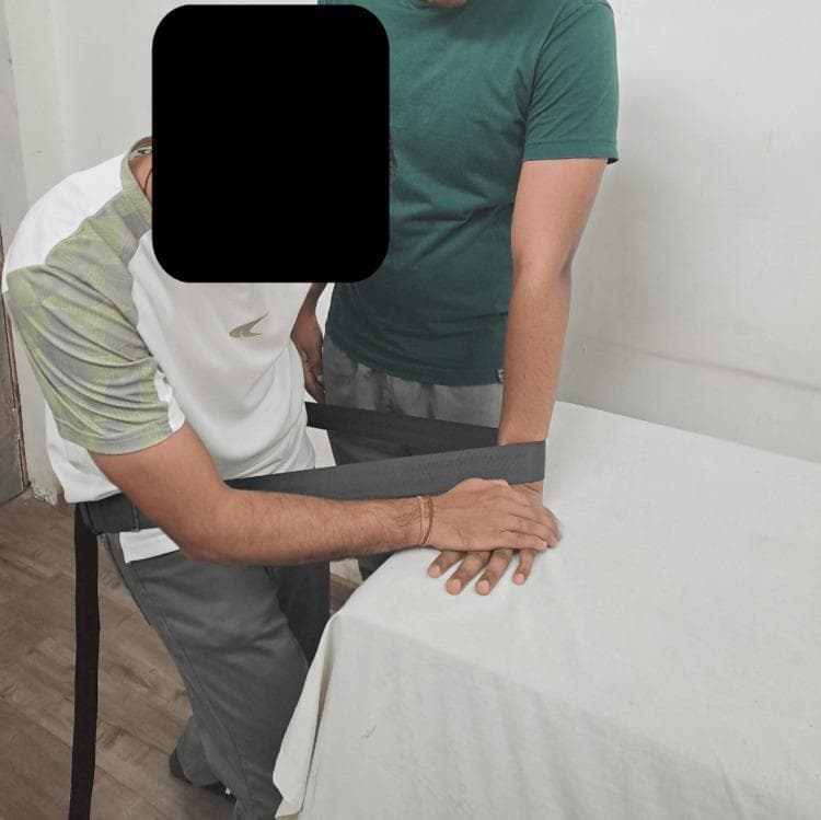 A clinical photograph illustrating a manual therapy technique for the wrist joint, specifically a weight-bearing lateral (ulnar) glide. The image depicts a physical therapist and a patient in a clinical setting. The patient stands with their forearm vertically oriented and the hand resting flat on a treatment table, simulating a weight-bearing position. The therapist is positioned to the left, utilizing a gray Mulligan belt (mobilization belt) looped around their own waist and the patient's distal forearm/wrist. This setup allows the therapist to use their body weight to provide a sustained lateral mobilization force. The therapist's hands are placed over the patient's wrist and hand to stabilize the joint and guide the glide direction. This technique is part of the 'Mobilization with Movement' (MWM) concept, typically used to improve range of motion and reduce pain in patients with musculoskeletal wrist dysfunction. The focus is on the biomechanical alignment and the application of manual force through specialized orthopedic equipment.