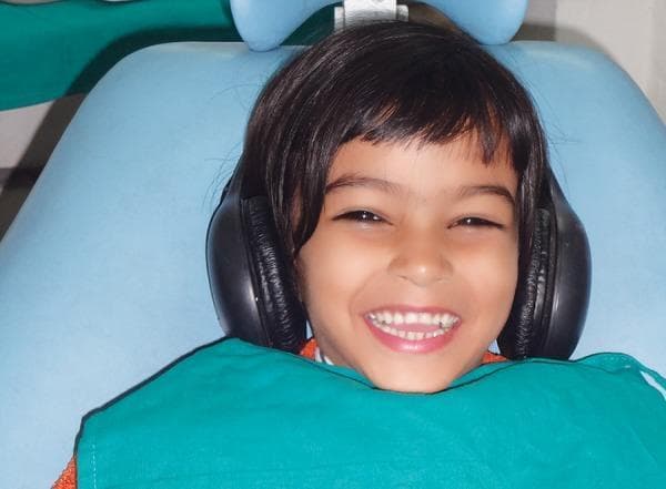 This clinical photograph illustrates a non-pharmacological behavior management technique used in pediatric dentistry. A young pediatric patient is shown in a dental chair, wearing large, over-the-ear black noise-canceling headphones. The patient displays a wide, smiling expression with visible upper and lower dentition and slightly squinted eyes, suggesting a positive emotional state and reduced anxiety. The child is draped with a green patient bib and positioned against the blue headrest of the dental unit. This image demonstrates the clinical application of 'audio analgesia' or music distraction to mitigate dental fear and distress during procedures such as tooth extractions. The target audience for this content includes dental students and pediatric practitioners exploring distraction therapy and its impact on physiological parameters like pulse rate and oxygen saturation during clinical interventions.