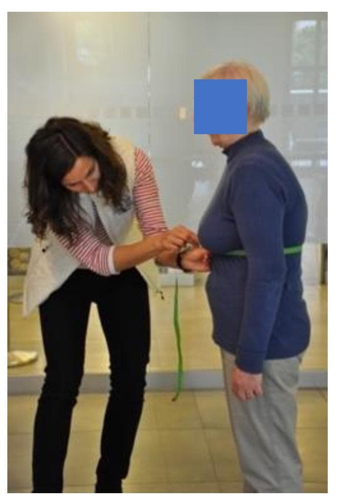 A clinical photograph demonstrating a circumferential chest expansion measurement, a physical assessment used to evaluate chest wall mobility and respiratory function. A clinician on the left is using a flexible green measuring tape to determine the thoracic circumference of an elderly patient. The tape is positioned horizontally around the patient's torso at the level of the xiphoid process of the sternum. This clinical procedure typically involves recording the difference in circumference between maximal inspiration and maximal expiration to assess lung excursion and rib cage flexibility. The measurement is relevant in the context of pulmonary rehabilitation for chronic respiratory diseases and the monitoring of conditions such as ankylosing spondylitis. The patient is shown standing in thin, close-fitting clothing with arms relaxed at the sides to ensure measurement accuracy.