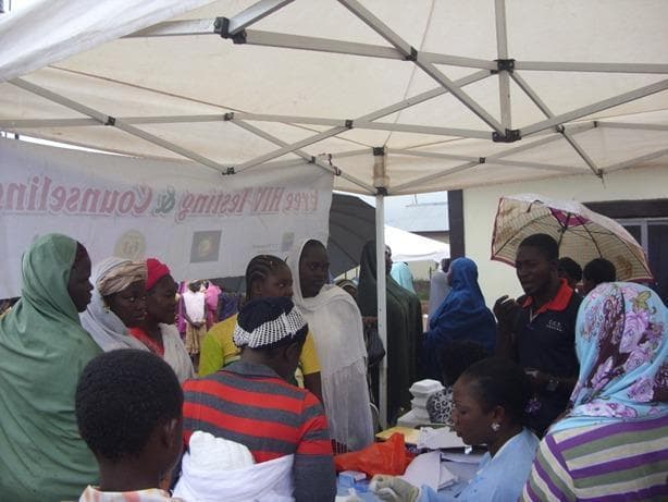 This clinical photograph captures a community-based medical outreach event focused on HIV testing and counseling in a low-resource or rural setting. The scene is organized under a temporary white canopy tent, providing a designated area for medical services. A prominent banner displays the text 'Free HIV Testing & Counseling,' indicating the primary public health objective. A healthcare worker, identifiable by blue clinical attire and protective gloves, is seated at a table covered with medical papers and testing materials, actively engaging with community members. The demographic depicted includes a diverse group of adults, both male and female, dressed in a variety of traditional head coverings and casual clothing, suggesting a culturally integrated approach to healthcare delivery. The setting emphasizes the concept of 'commonization' of HIV services by bringing diagnostic and counseling resources directly into hard-to-reach communities to improve accessibility and universal health coverage. The presence of individuals waiting in proximity to the testing station illustrates provider-initiated counseling and testing (PITC) in an informal, community-integrated environment.