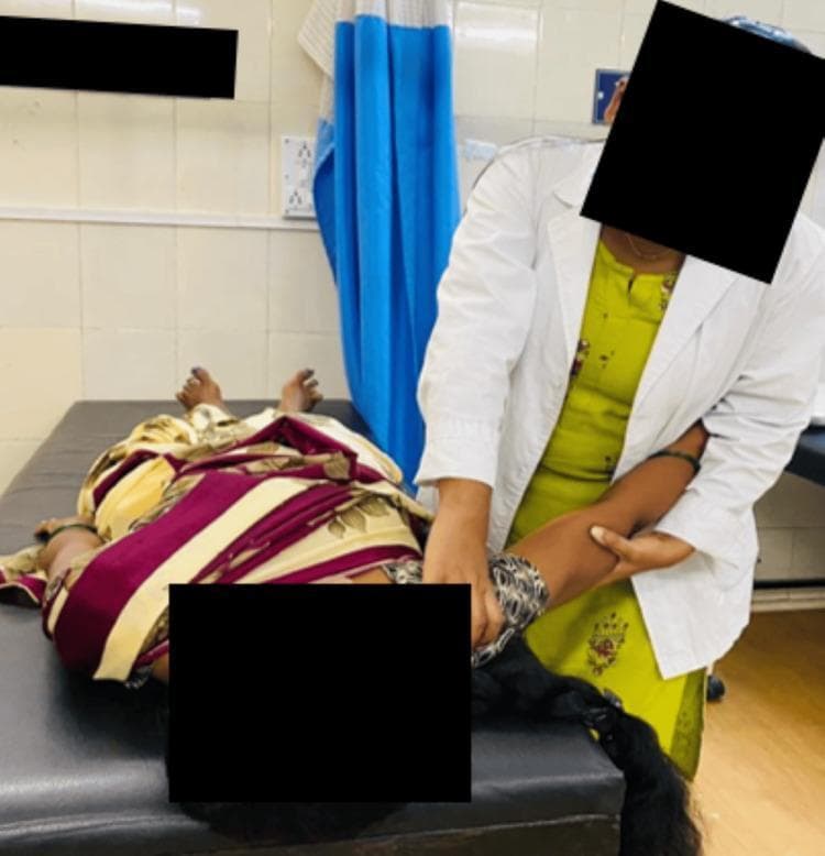 A clinical photograph depicting a manual therapy technique for the shoulder. A patient is positioned supine on a medical treatment plinth, with a physiotherapy practitioner standing at the bedside. The practitioner is performing a Maitland mobilization for the glenohumeral joint, specifically a caudal (inferior) glide in the resting position. One of the practitioner's hands is positioned near the patient's axilla to provide stabilization, while the other hand grasps the proximal humerus to apply a downward force. This orthopedic manual therapy intervention is commonly used in physical rehabilitation to improve range of motion and decrease pain in patients with conditions such as adhesive capsulitis or rotator cuff tears. The setting is a clinical environment with tiled walls and privacy curtains. The image serves as an educational demonstration of therapeutic hand placement and mobilization vectors for the upper extremity.