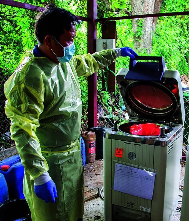 This clinical photograph depicts a health worker engaged in medical waste management, specifically the sterilization of infectious materials. The worker is outfitted in essential Personal Protective Equipment (PPE), including a long-sleeved, light yellow fluid-resistant gown, blue nitrile gloves, and a standard light blue surgical face mask. The healthcare professional is operating a vertical autoclave (steam sterilizer) used for decontamination. Inside the metallic chamber of the autoclave, a red biohazard bag is visible, signifying regulated medical waste such as COVID-19 related materials. The autoclave unit features a front panel with a pressure gauge, a biohazard warning label, and an attached log sheet for tracking sterilization cycles and compliance. This image illustrates critical infection control protocols, biosafety standards, and the procedural steps for medical waste disposal in a clinical or laboratory setting.