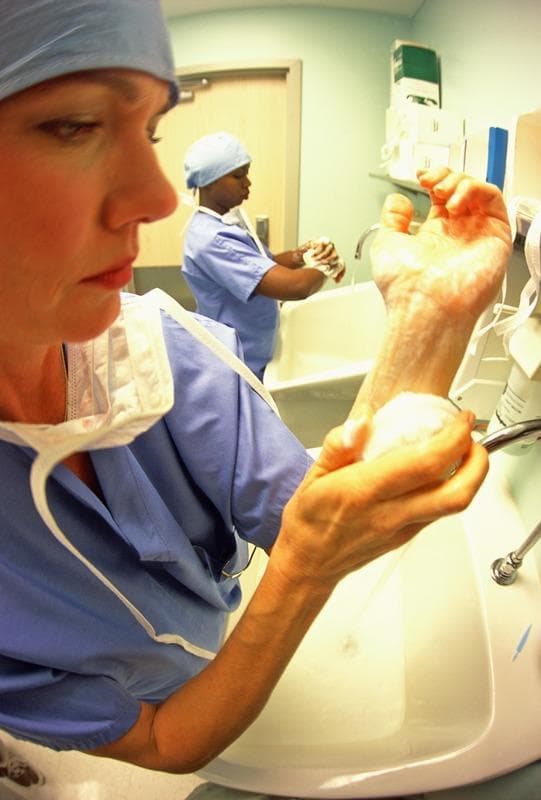This clinical photograph illustrates the essential medical practice of preoperative surgical hand scrubbing to ensure infection control and hospital hygiene. In the foreground, a healthcare professional is seen meticulously lathering their hands and distal forearms with an antimicrobial soap at a surgical sink. The individual is wearing standard operating room attire, including a blue surgical gown, a matching bouffant cap, and a white surgical mask draped around the neck. A second medical professional in similar surgical scrub attire is visible in the background, also engaged in hand washing at a separate station. The environment features standard medical fixtures, including deep white surgical sinks, chrome gooseneck faucets, and wall-mounted soap or paper towel dispensers. This image serves as an educational resource for demonstrating aseptic techniques, the prevention of nosocomial infections, and the rigorous hygiene protocols required for hospital staff before entering a sterile field.