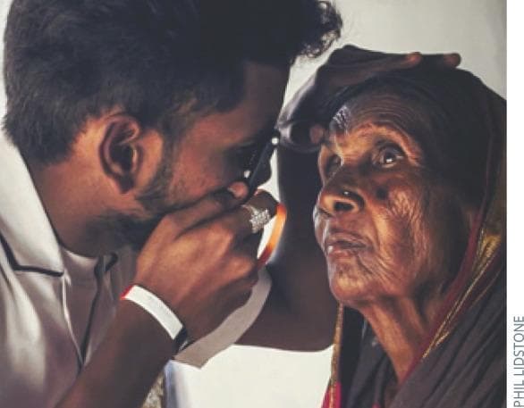 A clinical photograph illustrating the proper technique for direct ophthalmoscopy in a community health or low-resource setting. A healthcare provider is shown in close proximity to an elderly female patient with deeply wrinkled skin and a pigmented fundus. The provider is demonstrating the correct 'flight path' for visualizing the optic nerve, positioned on the horizontal plane at approximately 15 degrees temporal to the patient's right eye. The clinician holds a direct ophthalmoscope (specifically an Arclight device) in his right hand, using his right eye to examine the patient's right eye. His left hand is placed gently on the patient's forehead to stabilize the head and elevate the upper eyelid if necessary. The educational focus is on the physical positioning, equipment handling, and anatomical approach required to bring the optic disc and retinal vessels into view during a funduscopic examination. Relevant clinical concepts include screening for cataract, glaucoma, and macular disease through direct visualization of the posterior segment.