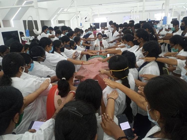 A clinical photograph depicting a large group of Phase I MBBS medical students participating in a formal 'Donor Oath' ceremony within an anatomy laboratory setting. The students, dressed in professional white coats and protective face masks, are gathered in a dense circle around a central dissection table covered with a reddish-pink cloth, likely containing a human cadaver. The individuals are performing a synchronized gesture, extending their right arms horizontally over the table, symbolizing their commitment to medical ethics, bioethics, and the respectful treatment of anatomical donors. The background shows specialized medical education infrastructure, including overhead surgical lighting and laboratory monitors. This image illustrates the integration of the Attitude, Ethics, and Communication (AETCOM) module in medical curricula, specifically focusing on humanizing anatomy education and instilling the principles of beneficence and non-maleficence in early undergraduate medical training.