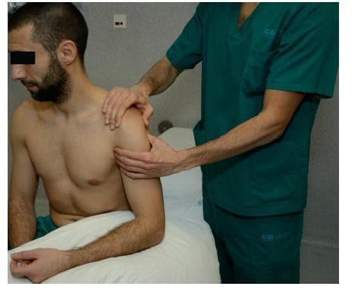 A clinical photograph demonstrating the performance of the Anterior Drawer Test of the shoulder, a physical examination maneuver used to assess glenohumeral joint instability. The image shows a shirtless male patient seated upright on an examination table with his right arm relaxed and the hand supported by a pillow. An examiner in teal scrubs is positioned behind and to the side of the patient. The examiner's left hand is used for stabilization, with the thumb placed on the coracoid process and the fingers spanning the scapular spine to fixate the scapula. The examiner's right hand grasps the proximal humerus, ready to apply an anteriorly directed force to provoke translation of the humeral head relative to the glenoid fossa. This procedural image illustrates the proper hand placement and patient positioning required to evaluate the integrity of the anterior capsulolabral structures and the degree of shoulder laxity.