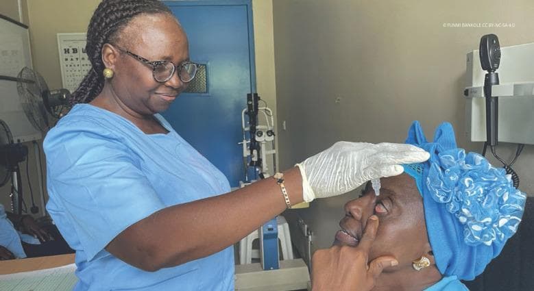 A clinical photograph depicting the administration of ophthalmic medication in a clinical setting. The image shows a healthcare professional, wearing blue scrubs and white disposable gloves, instilling eye drops into a patient's eye. The patient is reclined and assisting the procedure by gently retracting the lower eyelid with their finger to expose the conjunctival sac. A clear droplet is visible descending from a plastic dropper bottle toward the ocular surface. The background features ophthalmological equipment, including a slit lamp and an ophthalmoscope mounted on a wall bracket, indicating a professional eye care environment. This visual demonstrates standard clinical technique for topical medication delivery, patient positioning, and the use of personal protective equipment (PPE) in an outpatient ophthalmology or optometry clinic.