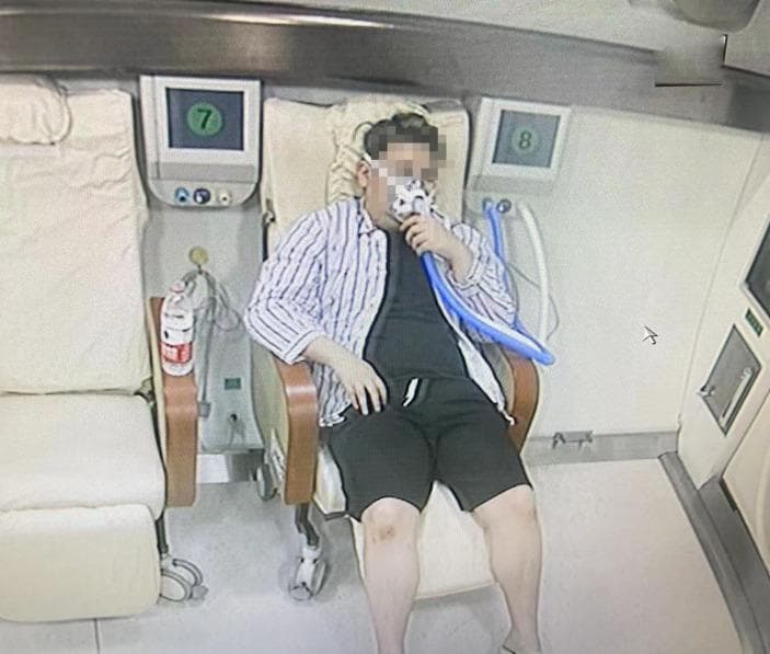 Clinical photograph of a patient receiving treatment inside a multi-place hyperbaric oxygen therapy (HBOT) chamber. The patient is shown in a seated, semi-reclined position wearing a tight-fitting oronasal oxygen mask connected to a blue corrugated delivery circuit. This setup facilitates the delivery of high-concentration oxygen under increased atmospheric pressure. The interior of the chamber features specialized medical seating, integrated digital monitoring screens labeled with station numbers, and wall-mounted gas delivery ports. Clinical context indicates this therapy is being utilized to manage peripheral neuropathy secondary to nitrous oxide (N2O) neurotoxicity. The image illustrates the application of hyperbaric medicine in treating neurological sequelae by increasing plasma oxygen solubility to counteract tissue hypoxia and potentially modulate cerebral blood flow and metabolic dysfunction caused by chemical inhalation.