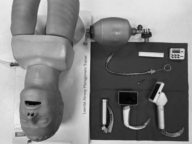 A high-angle clinical photograph displays standard and advanced airway management equipment for endotracheal intubation, positioned alongside a Laerdal Airway Management Trainer mannequin. The equipment includes three types of laryngoscopes: a conventional Macintosh-style laryngoscope with a metal handle and curved blade, and two video laryngoscopes featuring integrated digital screens and specialized curved blades for enhanced glottic visualization. Also present is a Bag Valve Mask (BVM) for manual ventilation. Centrally located is a clear cuffed endotracheal tube (ETT) with an internal stylet bent into a 'hockey-stick' configuration to facilitate insertion; the ETT includes a visible pilot balloon, connector, and depth markings. Ancillary procedural tools include a ruler for measurement and a digital timer for monitoring intubation time. This setup is typical for anesthesia training, emergency medicine simulation, or comparative studies between direct and video laryngoscopy techniques.
