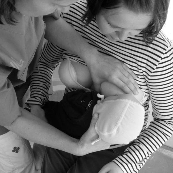 This clinical photograph captures a hands-on teaching interaction between a nurse and a mother within a neonatal or maternity care setting. The image demonstrates a breastfeeding support technique where the nurse is actively involved in positioning the infant for latching. The nurse, wearing professional medical scrubs, is positioned on the mother's left side. Her right hand is shown supporting the infant’s head and neck to maintain alignment, while her left hand is used to stabilize the mother’s breast to facilitate a proper latch. The mother is seated, wearing a striped shirt, and holds the infant close to her chest, focusing on the baby. The infant is wearing a bonnet and is swaddled, a common practice for maintaining warmth in neonatal care units. This visual illustrates the 'hands-on' approach to breastfeeding education, which is a clinical concept used to demonstrate physical positioning and latching techniques for new mothers, particularly in the context of preterm or sick infants requiring specialized support.