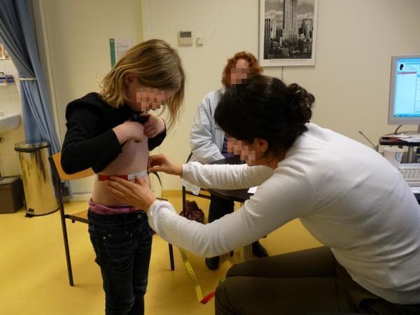 This clinical photograph depicts a general practitioner (GP) trainee performing an anthropometric measurement on a pediatric patient in a primary care setting. The professional is shown kneeling to maintain eye level with the standing child, using a flexible, non-stretch measuring tape to assess waist circumference. The child has appropriately lifted their shirt to expose the abdominal region, allowing for accurate tape placement midway between the lowest rib and the iliac crest. The procedure is conducted in a clinical environment, evidenced by the presence of an examination room sink, waste receptacle, medical privacy curtain, and a computer workstation. This image serves as an educational demonstration of standard physical examination techniques used in pediatrics to monitor nutritional status, assess metabolic risk, and calculate body mass index (BMI) correlates. The scene illustrates clinical best practices, including patient positioning and the use of standardized tools for longitudinal health tracking in general practice.
