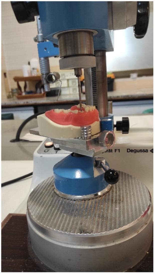 This clinical laboratory photograph illustrates the standardization of post space preparation in dental research using a precision milling machine. The setup features a Degussa F1 milling unit holding a dental model consisting of a maxillary arch with ivory-colored teeth and a pink gingival base. The model is secured onto a multi-axial adjustable platform, allowing for precise angulation and positioning. A high-speed drill head, mounted on a vertical support column, holds a #16 carbide round bur (44.5 mm height). The bur is shown in direct contact with the cervical/coronal portion of a specific tooth, simulating the vertical drilling required for uniform intracanal post space creation. Key visible components include the grooved base, the mechanical adjustment knobs for the milling arm, and the stable mounting of the specimen, which is essential for ensuring methodological consistency in endodontic and restorative dental studies. This procedure is critical for evaluating fracture resistance and the efficacy of different fiber post systems.