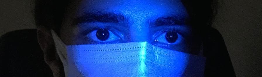 A clinical photograph of a patient undergoing an ophthalmic examination using slit lamp biomicroscopy. The image captures the periocular region of a male patient wearing a surgical mask, illuminated by a cobalt blue filter (450–500 nm wavelength) in a darkened environment. The primary focus is on the ocular surface and anterior segment. Despite the application of the specific blue light source often used to excite fluorescence (e.g., during fluorescein staining), there is a notable absence of ocular fluorescence in this specific view. The photograph serves as a negative control or comparison point in clinical investigations involving drug-induced ocular fluorescence, such as that associated with Favipiravir treatment. The educational significance lies in demonstrating standard slit lamp biomicroscopic examination lighting and the visual baseline for the ocular surface under cobalt blue light when no fluorescent substances or specific disease-related luminescence are present.
