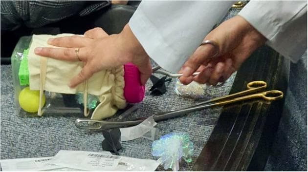 This clinical photograph captures a low-fidelity simulation of a Dilation and Evacuation (D&E) procedure, designed for medical education and physician training. The simulation model features a 'uterus' constructed from a clear plastic rectangular container, filled with various objects such as a yellow ball, green sponge, and iridescent mylar balls representing 'fetal parts.' The 'cervix' is simulated using a pink foam can holder secured to the container's opening, which is draped with beige fabric. A trainee's hands are visible, one stabilizing the model and the other operating a pair of silver-toned Bierer forceps to extract the contents through the simulated cervical canal. A pair of gold-handled surgical scissors lies on the surface nearby. This pedagogical tool is used to teach instrument handling, paracervical block techniques, and the tactile skills necessary for intrauterine procedures in obstetrics and gynecology. The setup illustrates an accessible, cost-effective method for clinical skills acquisition in reproductive health.