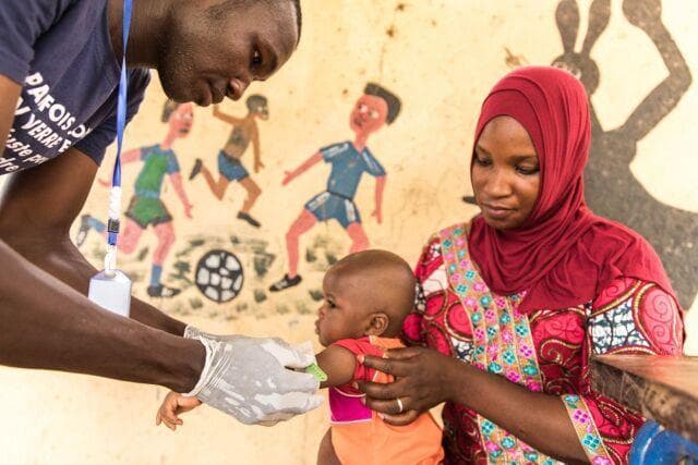 This clinical photograph depicts a Community Health Worker (CHW) in Mali performing a pediatric nutritional assessment. The healthcare worker, wearing protective gloves and a blue identification lanyard, is utilizing a Mid-Upper Arm Circumference (MUAC) tape—a color-coded screening tool—to measure the arm of an infant. The infant is being held by a caregiver, a woman dressed in a red hijab and patterned garment. This visual demonstrates primary healthcare delivery and growth monitoring in a resource-limited setting. The educational focus of the image is on the practical application of anthropometric measurements to screen for childhood malnutrition (wasting) at the community level. The setting is likely a village health post or mobile clinic, indicated by the hand-painted educational murals in the background. This image illustrates concepts related to global health, community-based primary care, and public health nutrition initiatives targeting rural populations.