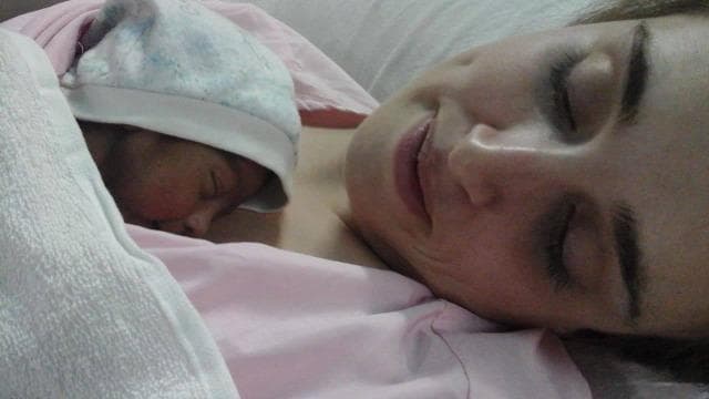 A clinical photograph illustrating Kangaroo Mother Care (KMC), also known as skin-to-skin contact, between an adult and a newborn infant. The image depicts the neonate positioned vertically and prone against the adult's bare chest, a key practice in neonatal care for temperature regulation and physiological stability. The infant is wearing a light-colored soft cap to prevent heat loss through the scalp and is partially covered by a pink blanket to maintain thermal insulation. Both the adult and the infant appear relaxed with eyes closed, demonstrating the bonding and calming effects of this intervention. This clinical practice is commonly utilized in maternal and child health settings, particularly in neonatal intensive care units (NICUs) and for low-birth-weight infants, to improve clinical outcomes such as breastfeeding success, neurodevelopmental stability, and infection prevention. The photograph highlights the human-centric approach to neonatal medicine and the integration of psychosocial support in clinical protocols.