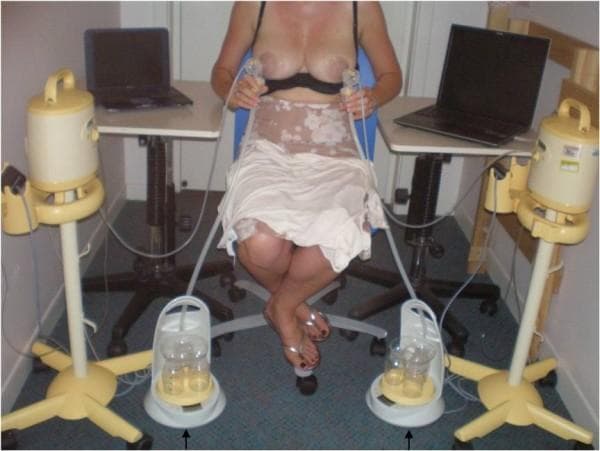 This procedural photograph illustrates a clinical research setup for simultaneous, bilateral milk flow rate measurement using the Showmilk technique. A seated participant is positioned between two hospital-grade electric breast pumps connected to bilateral breast shields. The experimental apparatus utilizes clear collecting tubes that transport expressed milk from the shields down to purpose-built continuous weight measurement devices (Showmilks) positioned on the floor. These weighing devices feature circular bases with platforms housing multiple collection bottles for fractional sampling. The system is integrated with two pressure transducers to monitor vacuum levels. Data from the weight balances and transducers are transmitted via USB interfaces to two notebook computers for real-time visualization and recording of milk ejection patterns. This setup is designed to evaluate milk ejection frequency, flow rate peaks (g/s), and synchronicity between breasts during expression, providing objective data on the physiological mechanisms of lactation and the milk ejection reflex.