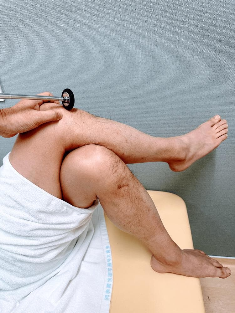 This clinical photograph demonstrates the assessment of the patellar tendon reflex (knee-jerk) in a patient positioned supine. The patient's legs are arranged with the right leg crossed over the left, placing the uppermost knee in a flexed position that naturally stretches the quadriceps tendon. An examiner’s hand is visible, stabilizing the knee while using a reflex hammer with a circular rubber head to strike the patellar ligament just distal to the patella. This procedure is used to evaluate the L3-L4 spinal nerve roots and the integrity of the femoral nerve. The image illustrates a practical modification for eliciting deep tendon reflexes when a patient is in a semi-recumbent or supine position on an examination table, emphasizing proper limb positioning and strike location for clinical diagnostic purposes.