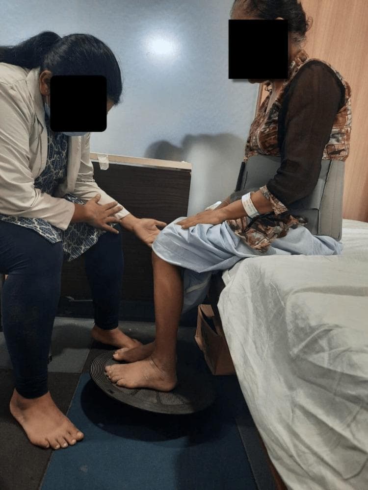 A clinical photograph capturing a postoperative physiotherapy session for a patient following lumbar spine surgery. The patient is seated at the edge of an examination bed, wearing a rigid lumbosacral orthosis (back brace) for spinal stability. The patient's bare feet are positioned on a black circular wobble board placed on a blue non-slip floor mat. A physiotherapist is positioned in a semi-kneeling stance to the patient's side, providing manual guidance and tactile cues at the patient's knee and arm to assist with balance. This setup illustrates a seated proprioceptive and neuromuscular retraining exercise, designed to improve core stability and lower extremity motor control while minimizing spinal load during early mobilization. The context suggests rehabilitation following L3-S1 decompression and fusion for spinal stenosis.