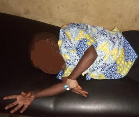 This clinical photograph displays a pediatric patient demonstrating classic carpopedal spasm, a clinical sign of hypocalcemia (Trousseau sign of latent tetany) often secondary to dyselectrolytemia. The child is shown in a recumbent position on an examination surface. The left hand and forearm are prominently featured, revealing a characteristic posture: flexion at the wrist and metacarpophalangeal (MCP) joints, with extension of the interphalangeal (IP) joints and adduction of the thumb ('obstetrician's hand' appearance). An intravenous (IV) catheter is visible on the left forearm, secured with medical tape and a hub, indicating ongoing medical intervention for fluid or electrolyte resuscitation. This visual serves as a primary diagnostic indicator for neuromuscular irritability and tetany, frequently associated with metabolic disturbances such as hypocalcemia, hypomagnesemia, or metabolic alkalosis.