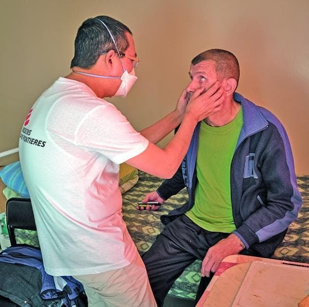 This clinical photograph depicts a physical examination during a patient consultation at a pulmonology and tuberculosis center. A healthcare professional, identified by 'Médecins Sans Frontières' branding on a white t-shirt, is performing a manual assessment of a seated patient's face. The clinician is wearing corrective glasses and a white surgical mask for infection control, which is essential in a respiratory health setting. The examination involves the clinician using both hands to palpate the patient's submandibular or malar region, while the patient looks upward, potentially allowing for the inspection of the conjunctivae for pallor or checking for lymphadenopathy. The setting is a clinical ward or residential treatment room, indicated by the bed and personal items. This image illustrates humanitarian medical aid, infectious disease screening (specifically tuberculosis monitoring), and the importance of personal protective equipment (PPE) during direct patient contact in respiratory specialty clinics.