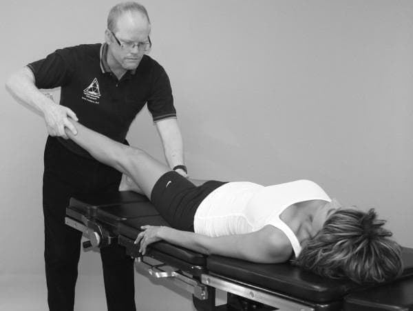 A clinical photograph in black and white demonstrating a Manual Muscle Test (MMT) for the tensor fasciae latae (TFL). The image depicts a patient in a supine position on a clinical examination table. The patient's right leg is extended and elevated at an angle, representing the test position for TFL assessment involving hip flexion, abduction, and internal rotation. A male clinical examiner stands at the foot of the table, applying manual resistance to the patient's distal lower leg near the ankle with both hands. The examiner is positioned with an upright posture, maintaining visual focus on the limb's response. The patient's head is turned away from the side being tested. This visual serves as an educational guide for physical therapy, chiropractic, or orthopedic assessment, illustrating proper practitioner hand placement, patient positioning, and the line of force required to isolate the tensor fasciae latae while minimizing synergist substitution during muscle strength evaluation.