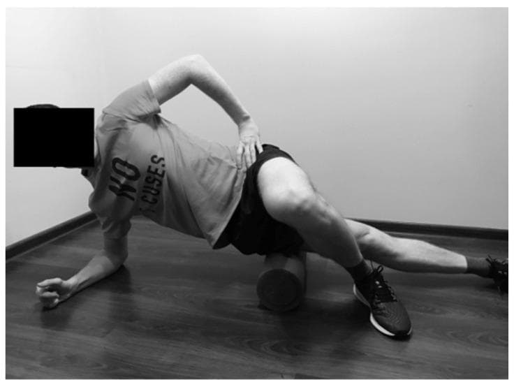 This clinical photograph demonstrates a self-myofascial release (SMFR) technique for the tensor fasciae latae (TFL) and iliotibial (IT) band. The individual is positioned in a lateral side-bridge posture on a wood-laminate floor. Support is maintained by the left forearm and the right foot, which is crossed over the lower leg and planted firmly on the ground to control pressure and movement. A high-density cylindrical foam roller is placed transversely under the lateral aspect of the left thigh, specifically between the iliac crest and the knee joint. The subject’s right hand is placed on the hip to stabilize the torso, which is slightly rotated anteriorly. This exercise is used in physical therapy and sports medicine to improve soft tissue extensibility, reduce myofascial hypertonicity, and manage conditions such as iliotibial band syndrome. The image illustrates the starting position for rolling along the lateral kinetic chain to address muscle tightness in the hip abductors.