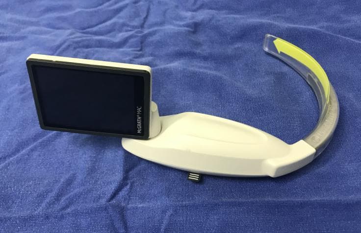 This clinical photograph displays a McGrath MAC video laryngoscope, an essential airway management tool used for endotracheal intubation. The device consists of a white, ergonomically contoured handle and a built-in rectangular LCD screen with a black bezel labeled 'McGrath® MAC'. Attached to the handle is a curved, Macintosh-style blade. The blade assembly features a metallic core encased in a clear, disposable transparent plastic sheath, which is characterized by a prominent yellow directional arrow pointing toward the distal tip. The device is shown resting on a blue surgical drape, highlighting its role in anesthesiology and emergency medicine. This visual is designed to educate medical professionals on the physical components and assembly of video laryngoscopy equipment used to improve laryngeal visualization during difficult airway scenarios.